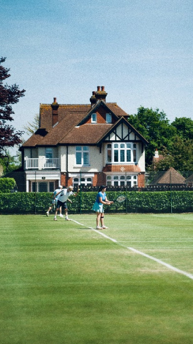 People playing tennis on grass courts. In the background a half-timbered house.